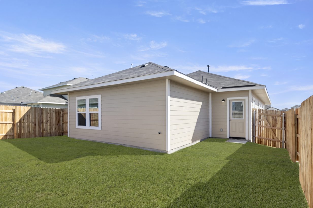 Image of the back exterior of a tan one story house with white trim, green grass back yard, a wooden fence and a blue sky in the background
