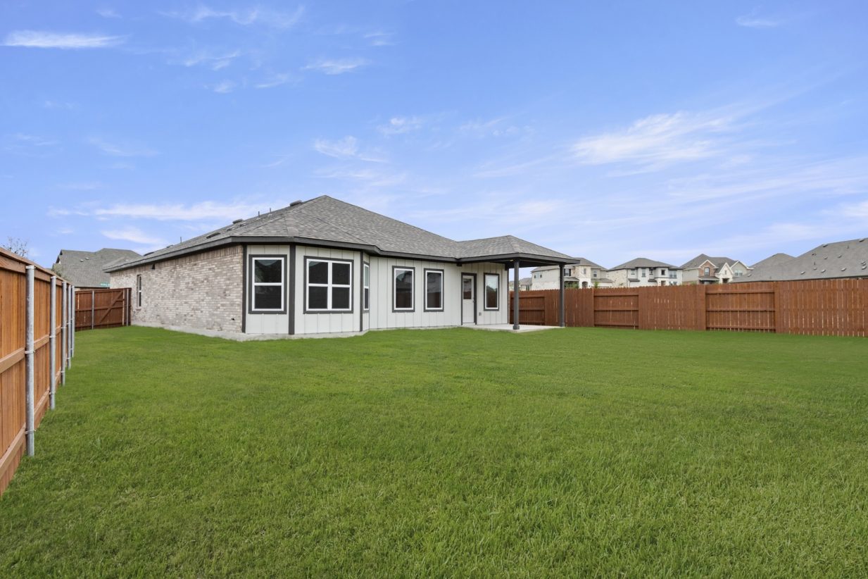 Image of a back exterior of a one story brick home with green grass and a brown wooded fence and a blue sky in the background