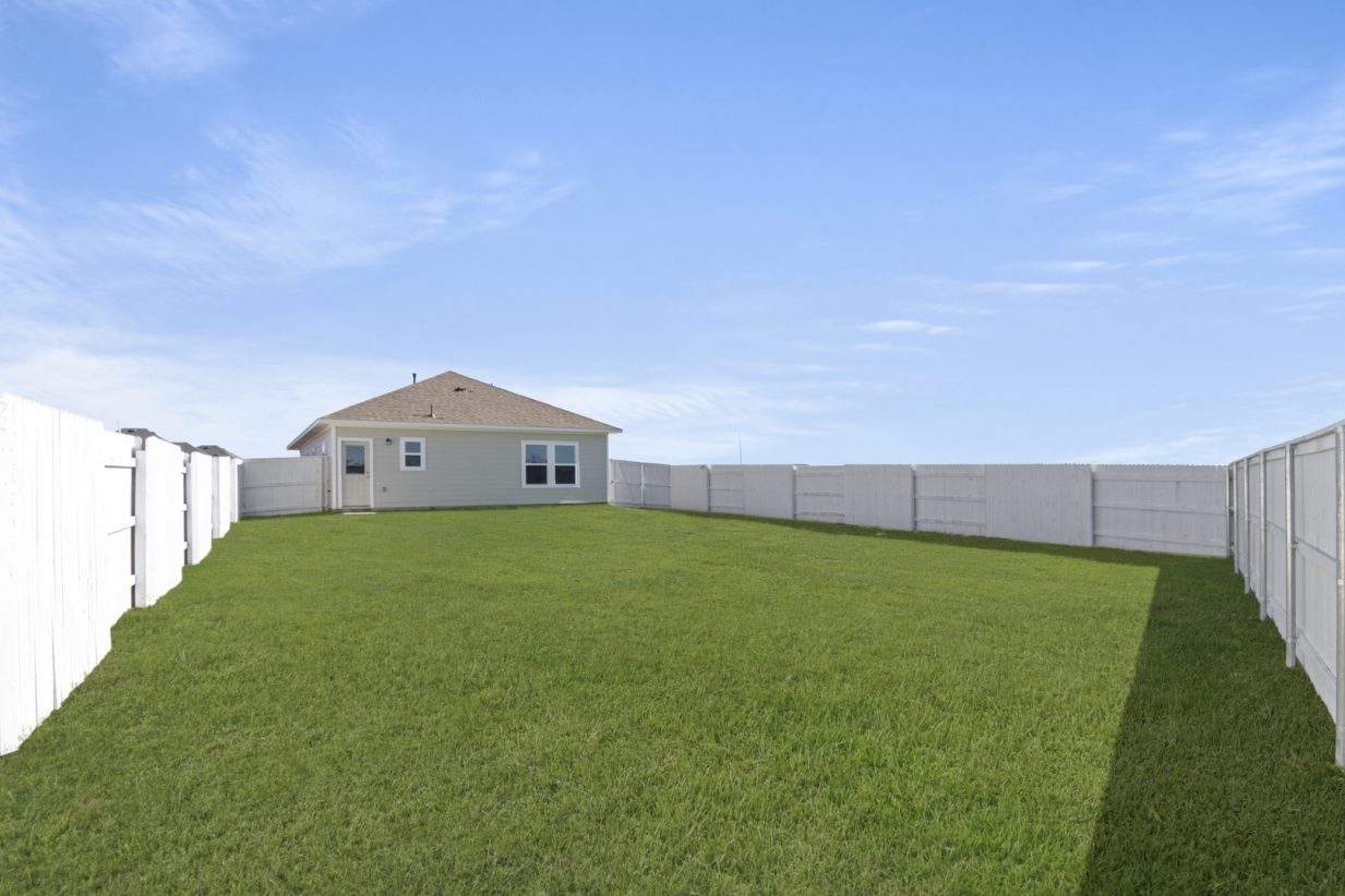 Image of a backyard exterior with a large green grass area and a white wooden fence and a blue sky in the background