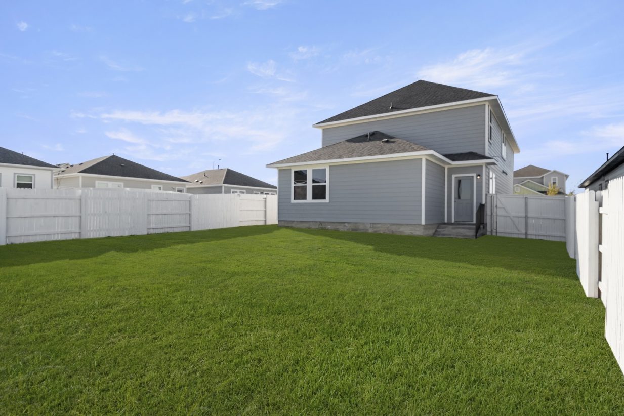 Image of the back exterior of a grey two story house with a large green grass backyard, a white wooden fence, and a blue sky.