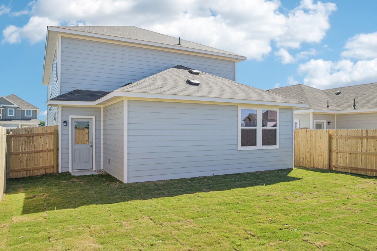 Image of a blue two story house back exterior with a green grass backyard, wooden fence and a blue sky in the background