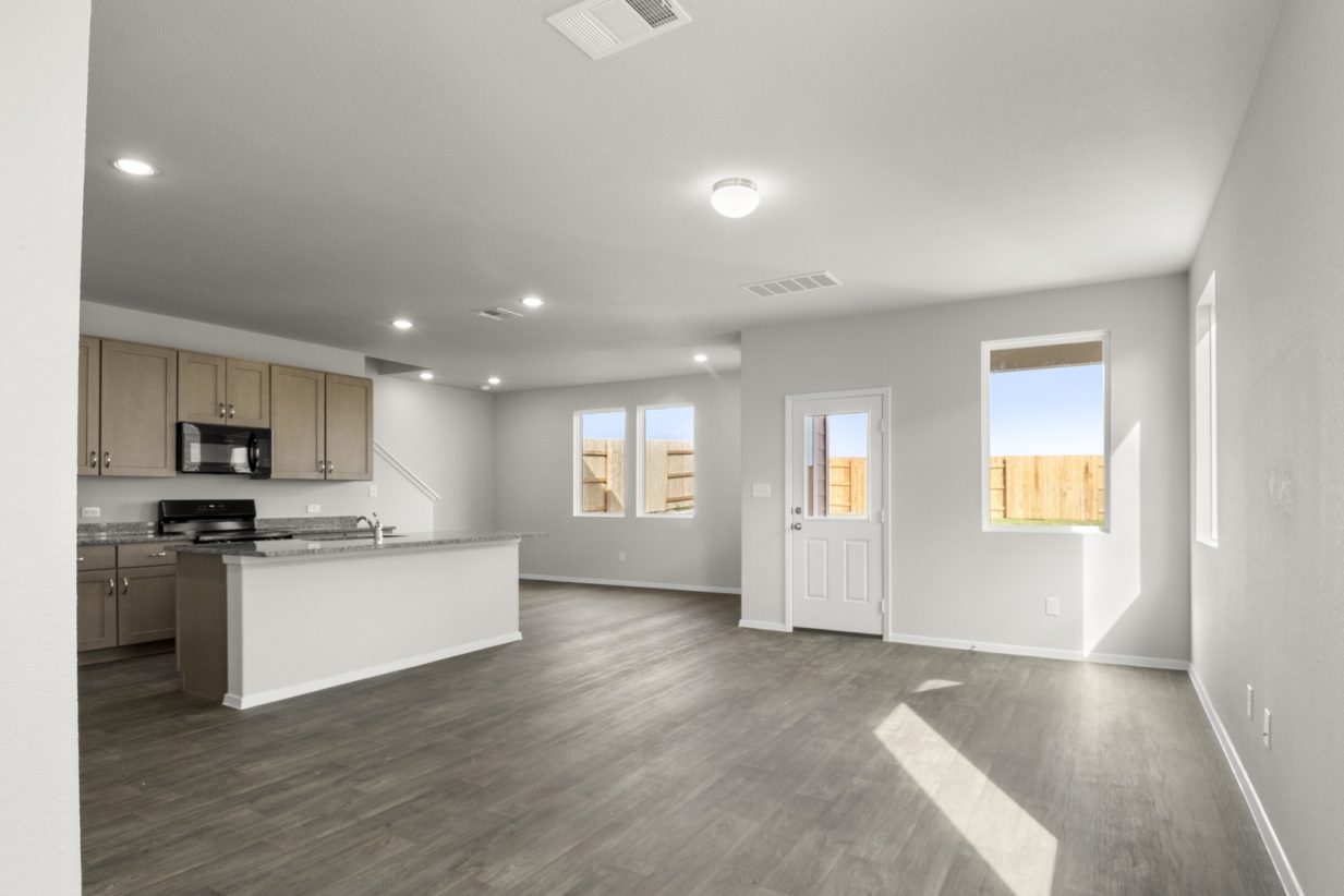Image of a living room with light grey walls, dark brown floors, windows and a kitchen in the distance