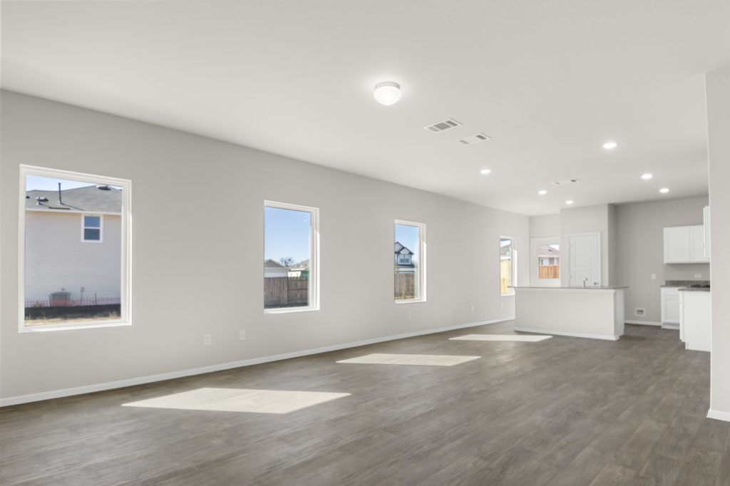 Image of a living room with cream walls, dark brown flooring, white trim, windows and a kitchen in the distance