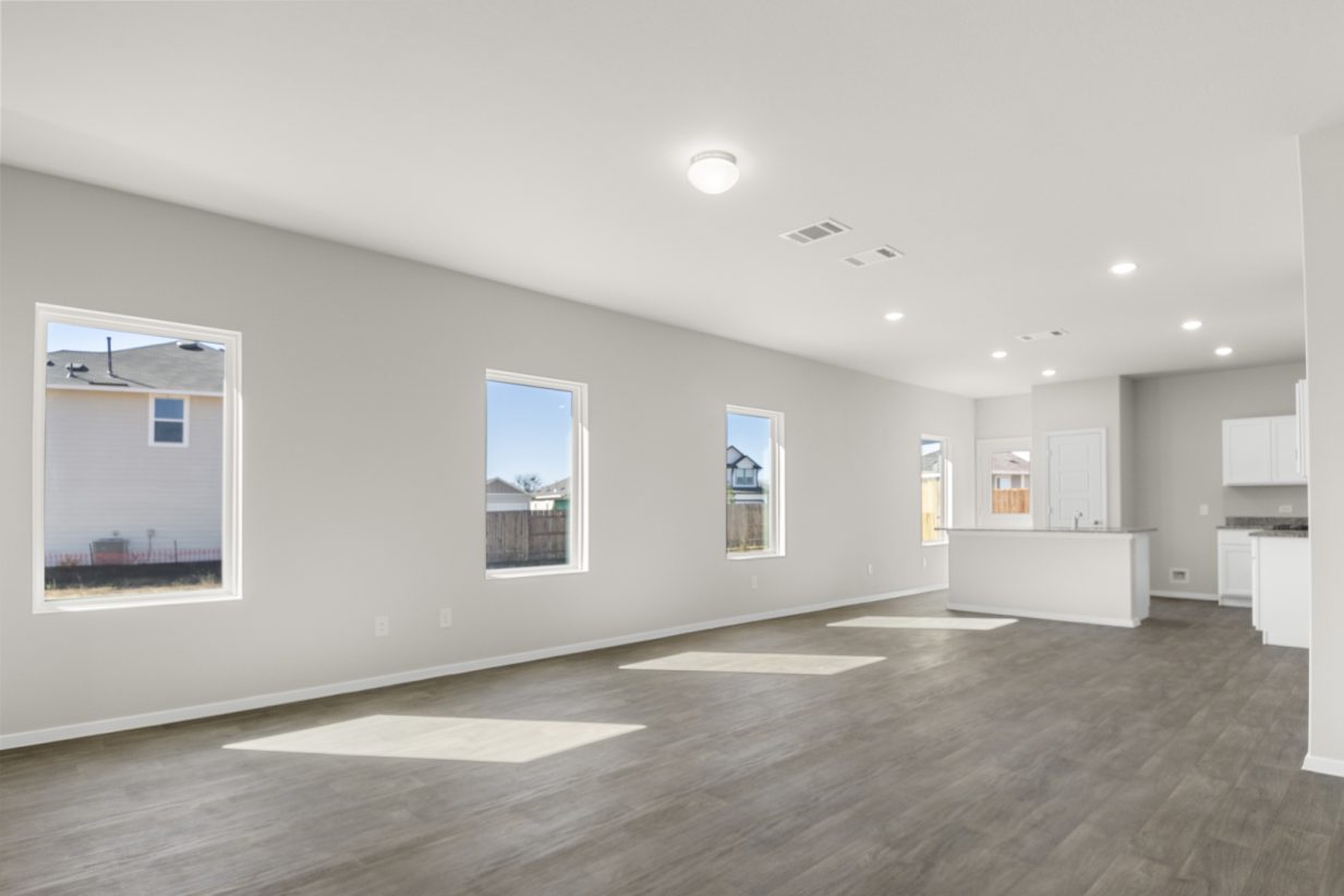 Image of a living room with cream walls, dark brown flooring, white trim, windows and a kitchen in the distance