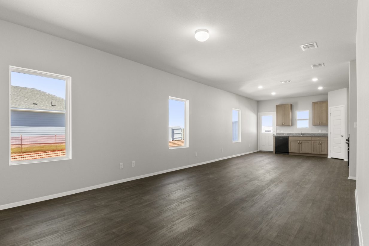 Image of a living room with vinyl flooring and light grey walls and two windows and a kitchen in the distance