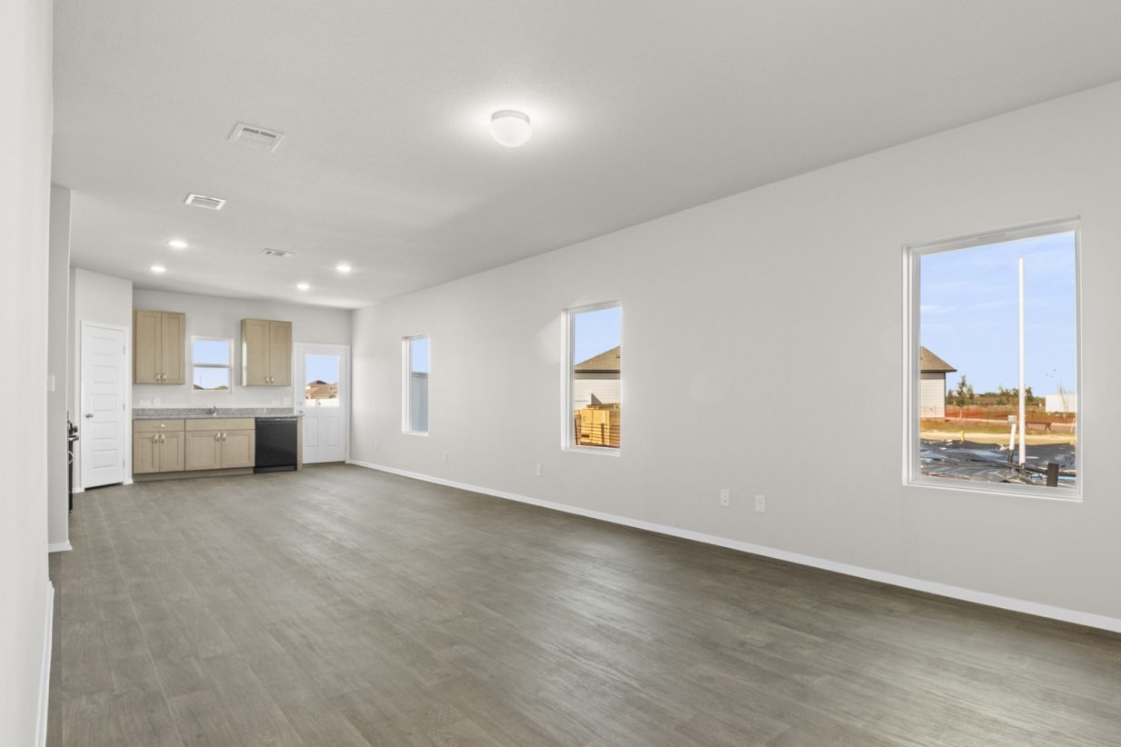 Image of a living room with dark vinyl flooring, light grey walls, windows and a kitchen in the distance