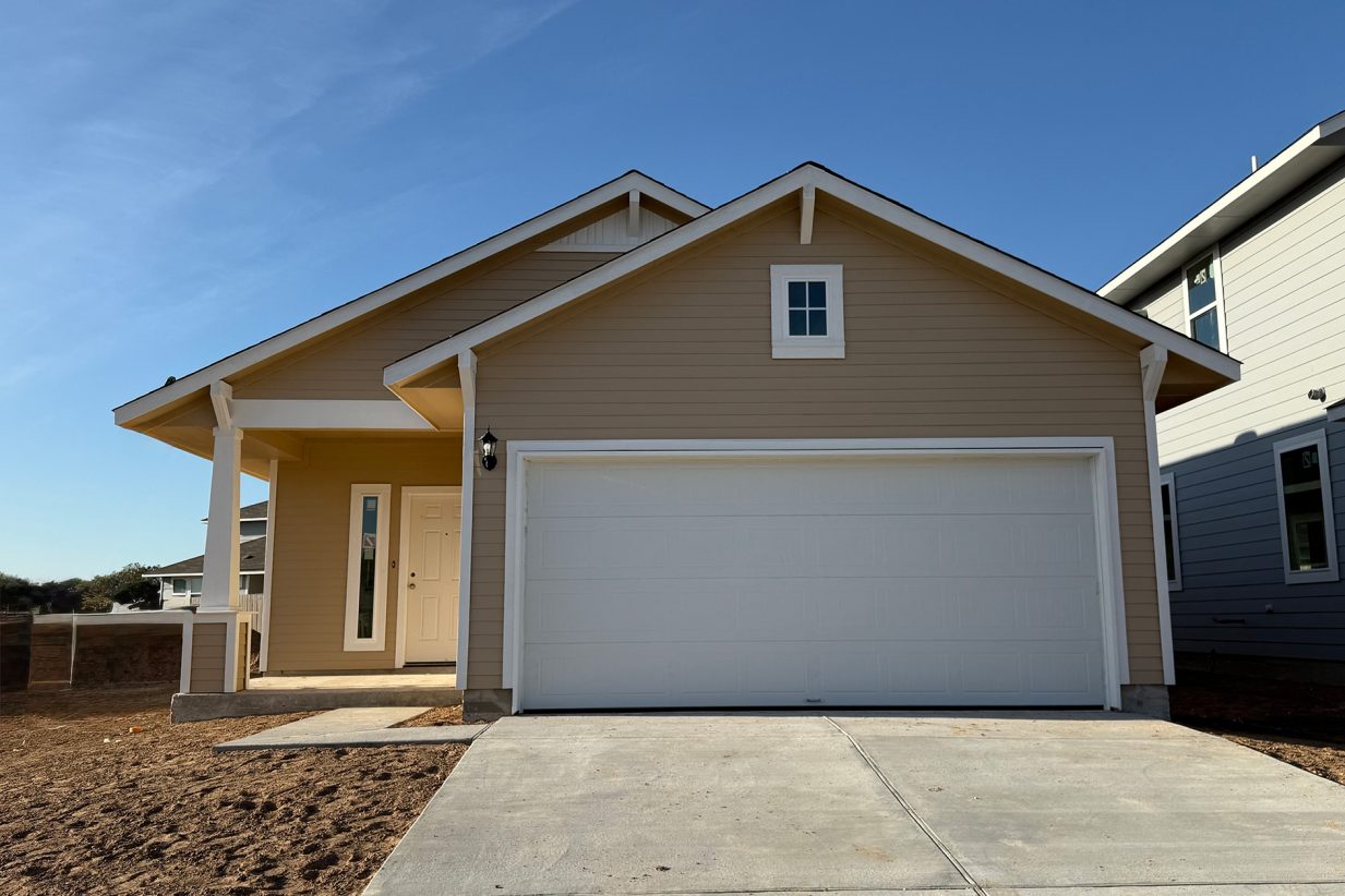 Image of a front exterior of a tan one story house with a white garage door with a cement driveway and a blue sky in the background