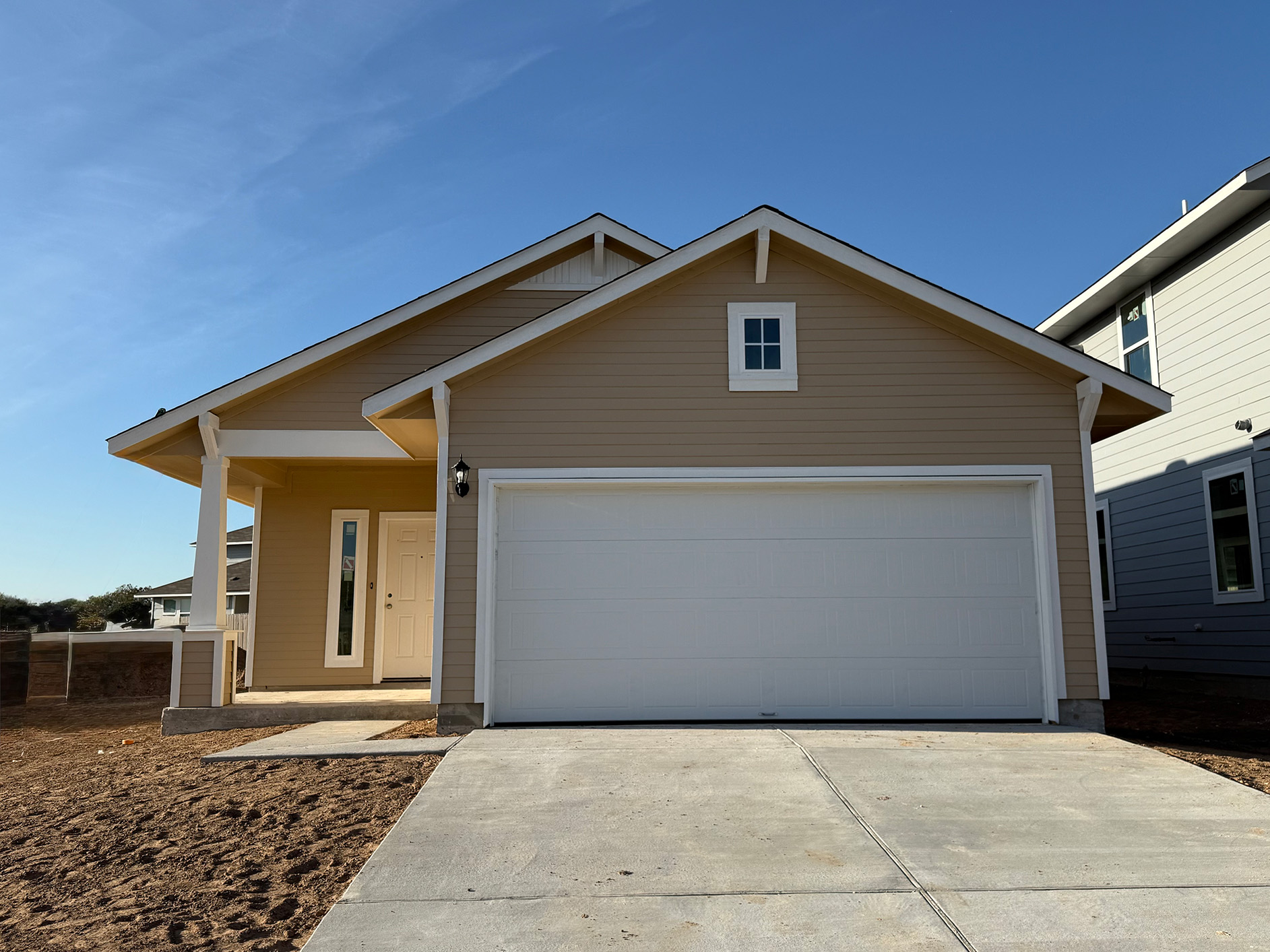 Image of a front exterior of a tan one story house with a white garage door with a cement driveway and a blue sky in the background