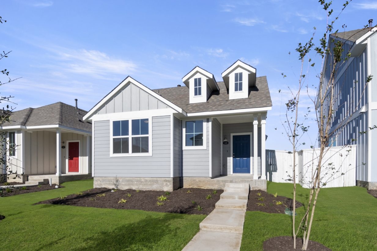 Image of the front exterior of a grey one story home with white trim, a blue front door, green grass and a blue sky in the background