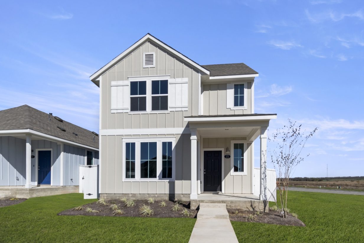Image of a tan two story house front exterior with a black front door, white trim, windows, a green grass front yard and a blue sky in the background