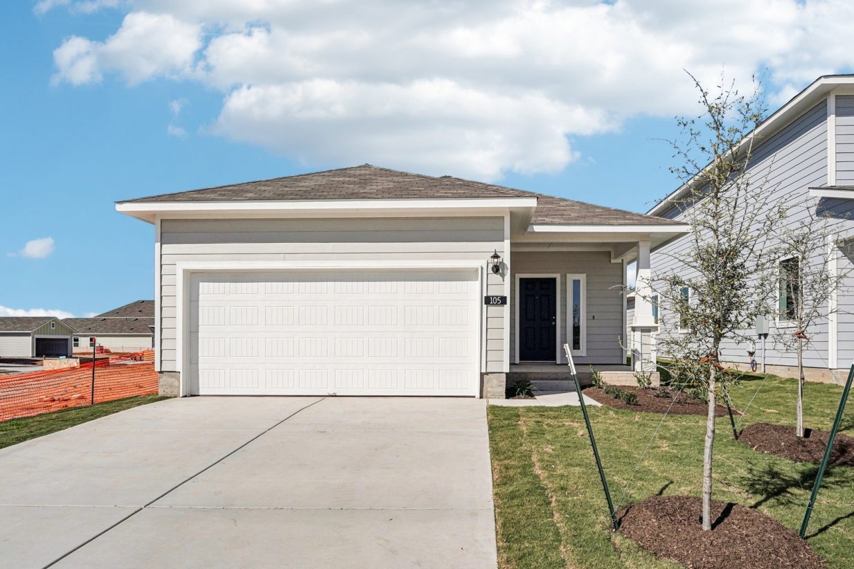 Image of a grey one story house with a white garage, cement driveway, a black front door and small trees