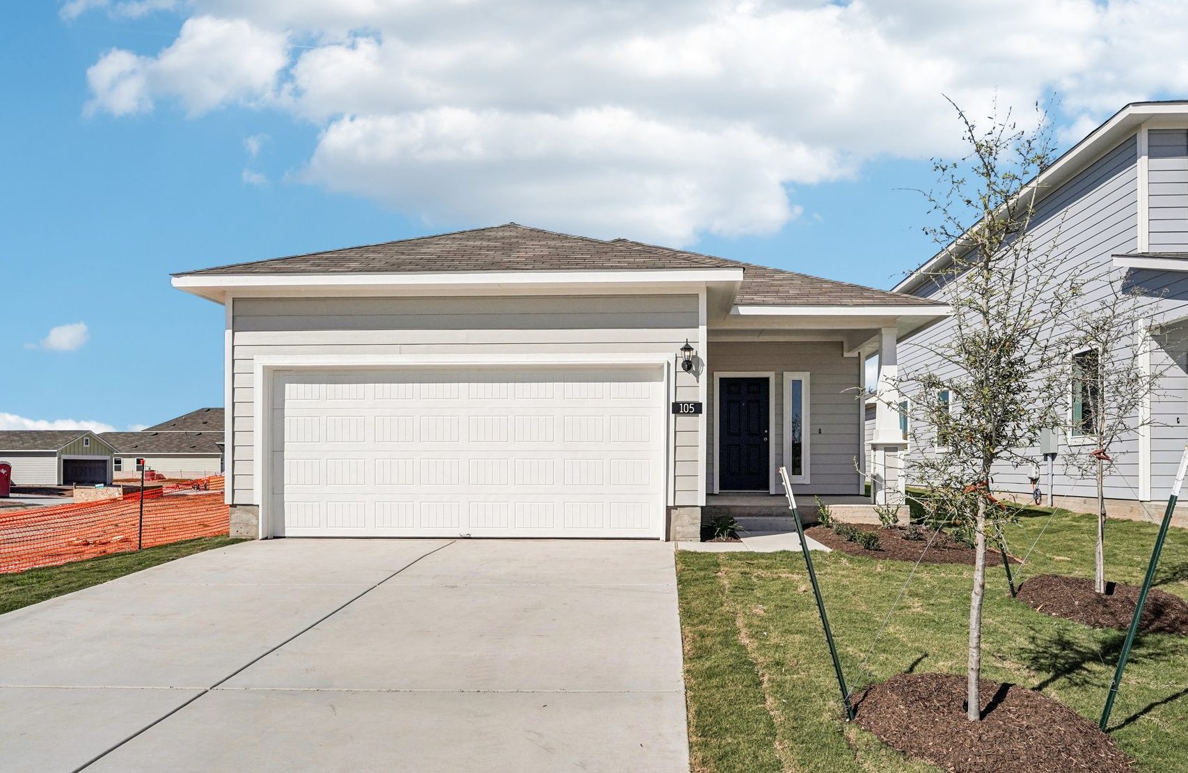 Image of a grey one story house with a white garage, cement driveway, a black front door and small trees