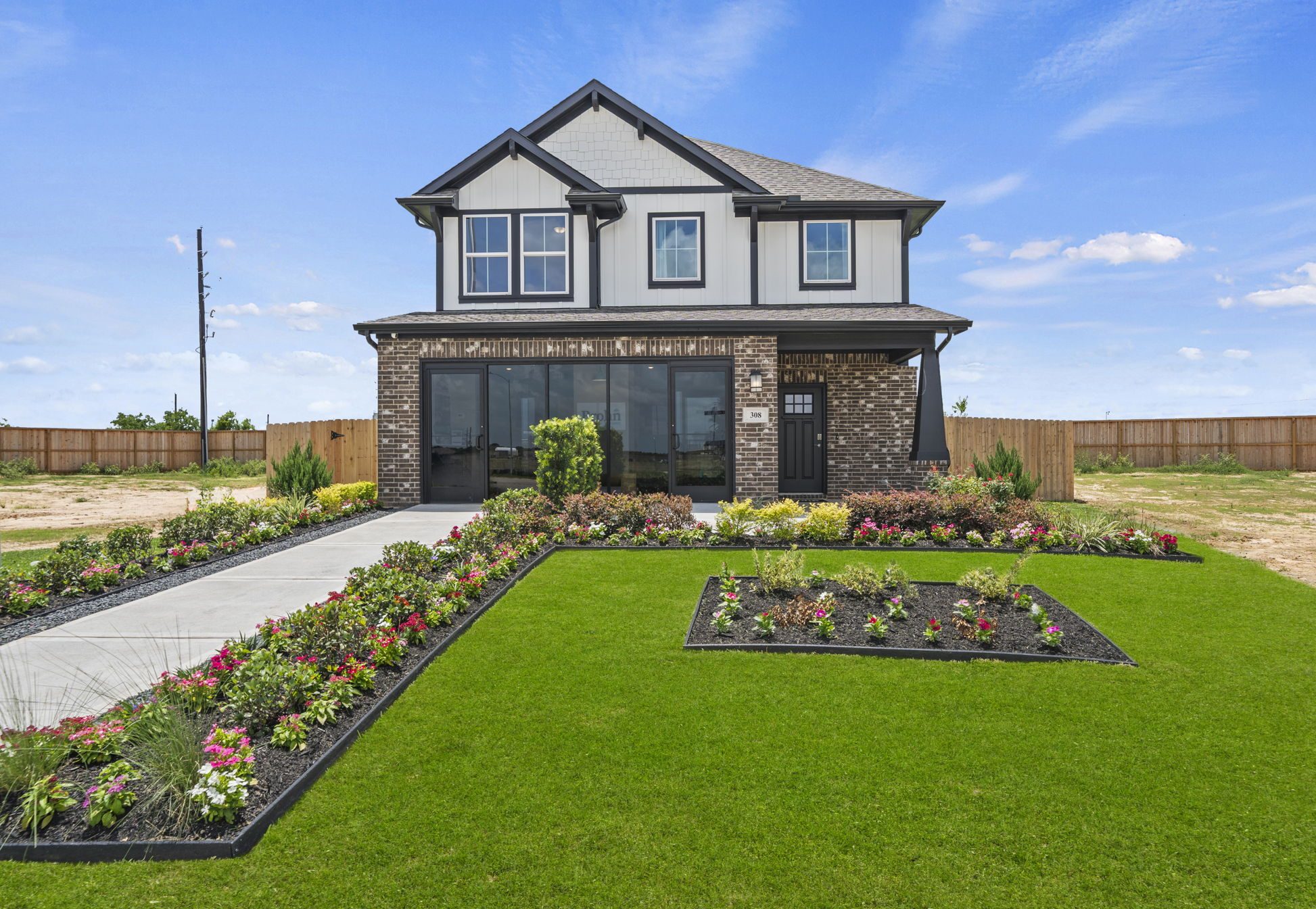 An exterior image of a two-story white house with black trim, accented by brown brick, with flower landscaping and green grass, on a concrete sidewalk, set against a blue sky background.