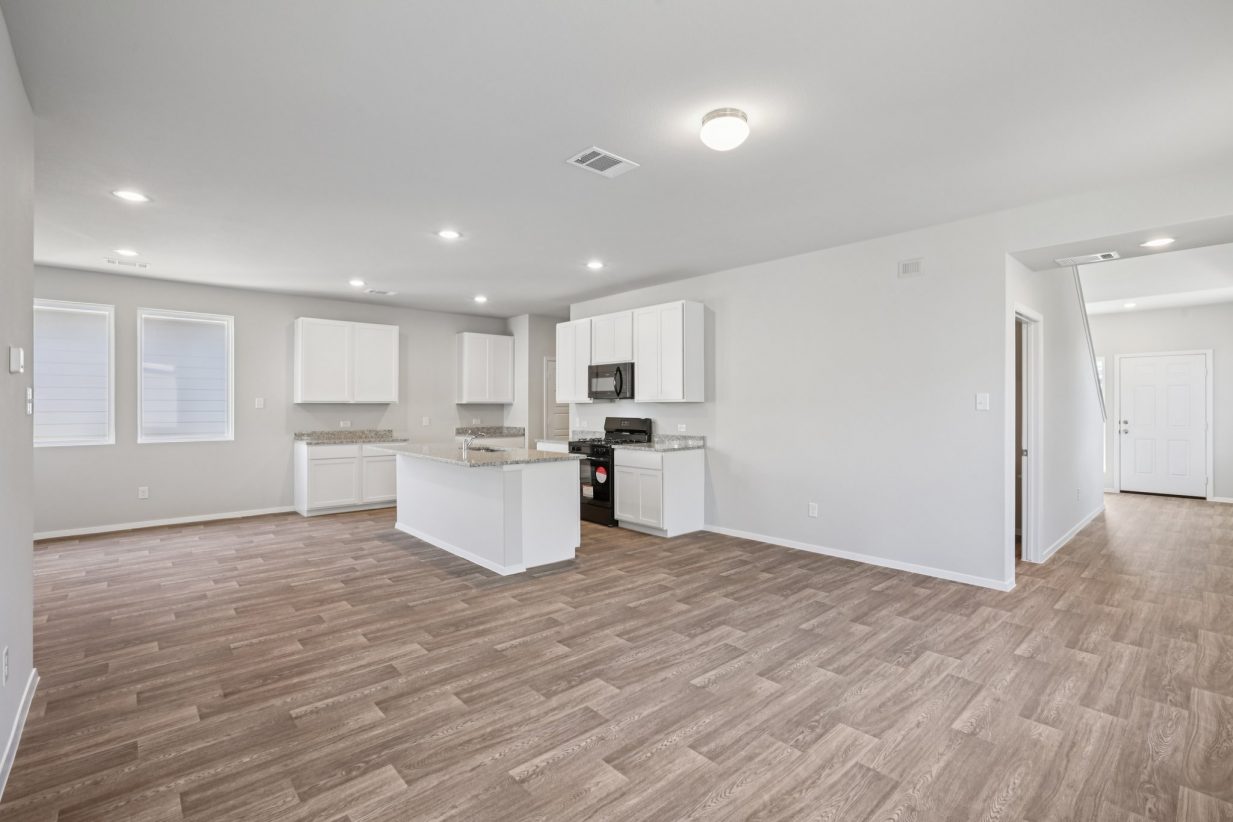 Image of an open living room with grey walls, light brown vinyl flooring, an open kitchen in the corner and a staircase to the left