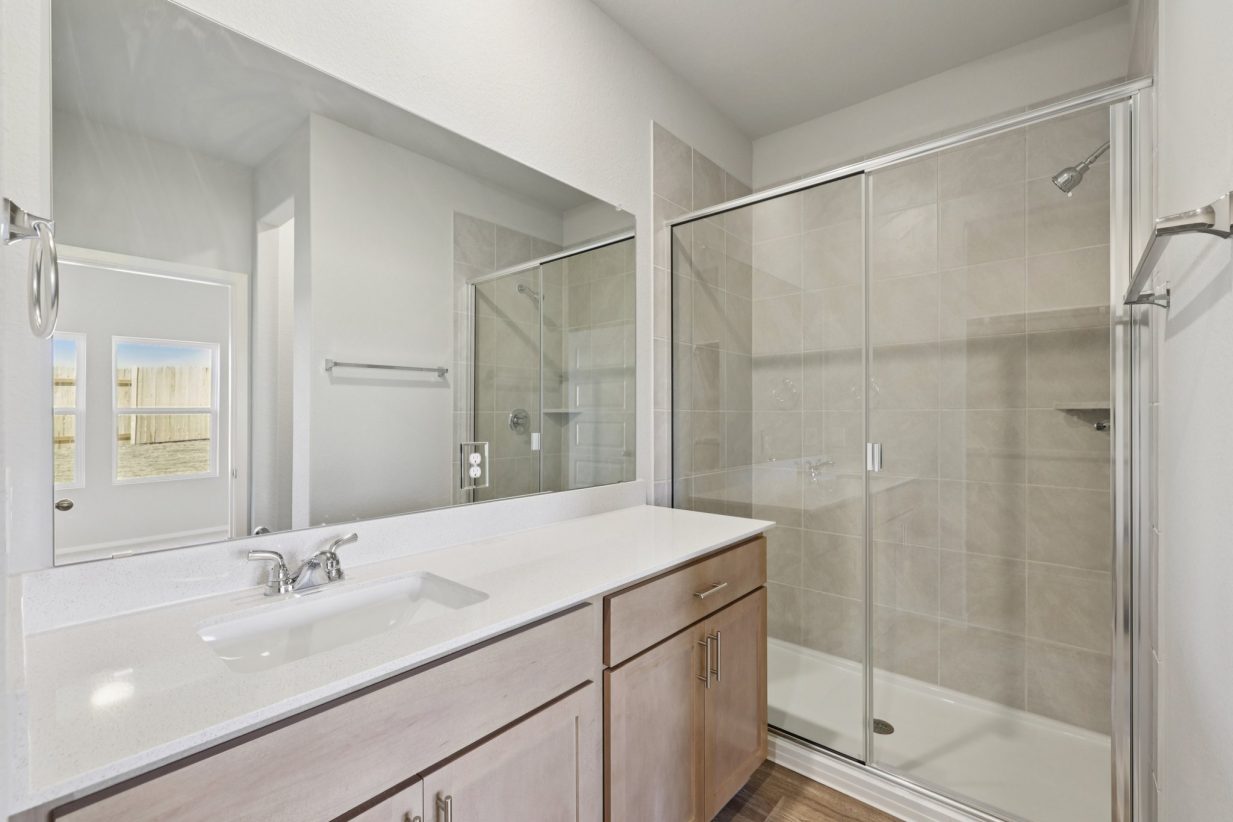 Image of a primary bathroom with a white dual vanity, light brown cabinets, a large mirror, and a standing shower