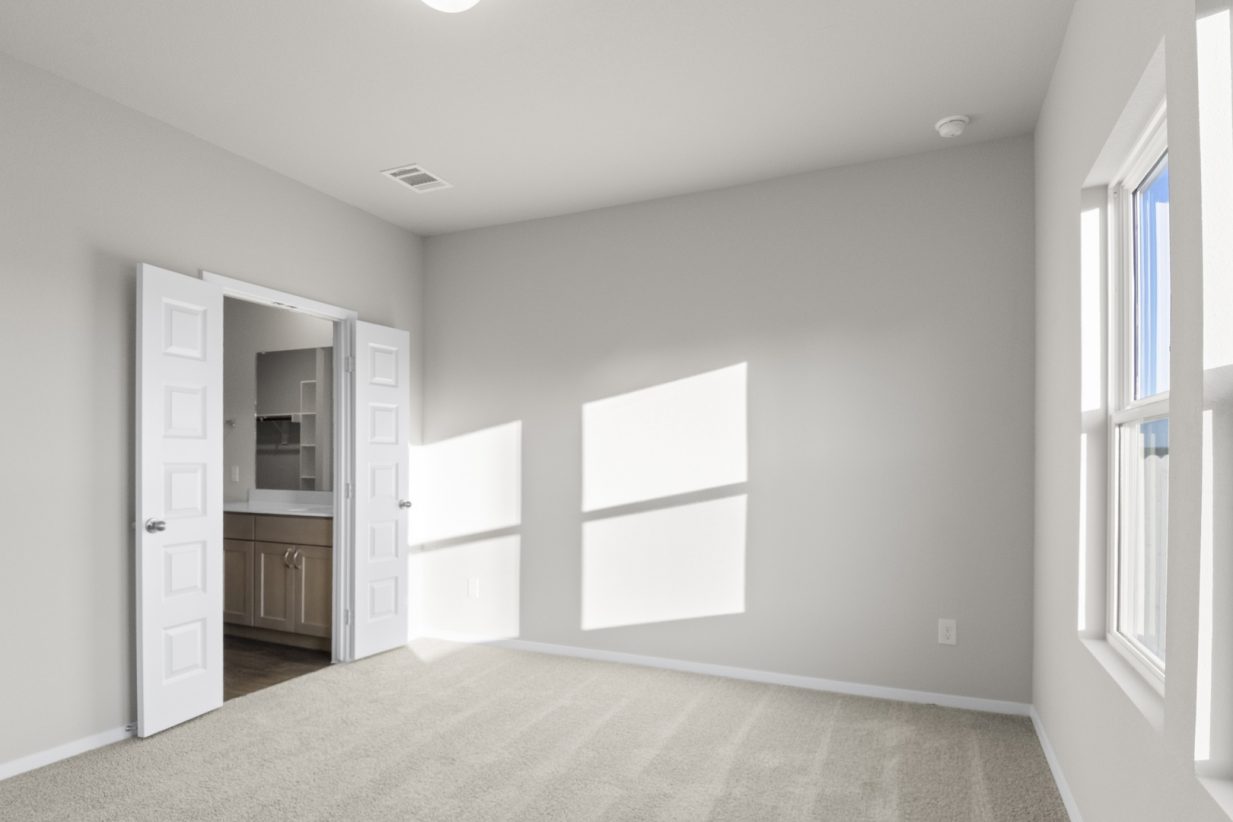 Image of a one story home bedroom with tan carpeting and light grey walls with two windows on the right and open door looking into en suite bathroom with vanity with white countertop and light brown cabinets