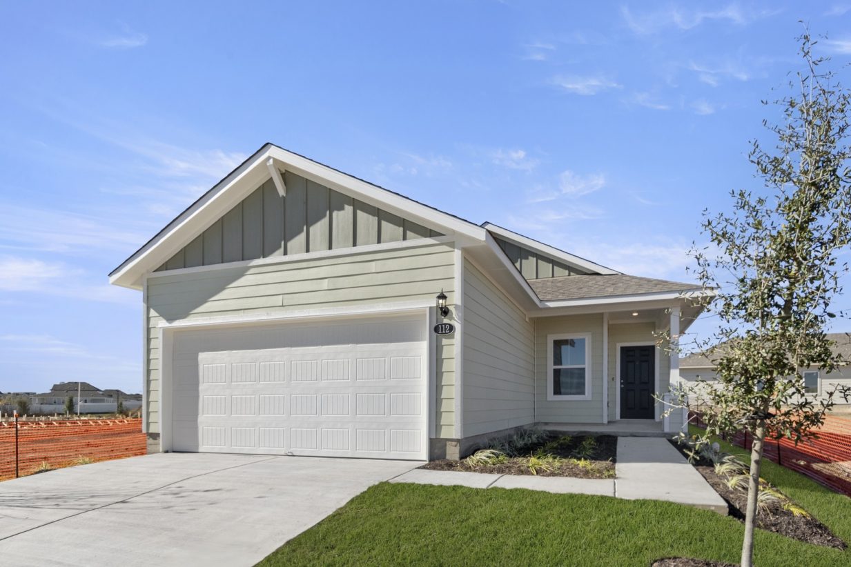 Image of a light green one story home front exterior with a white garage door, white trim, a black door, a cement driveway and a light blue sky in the background