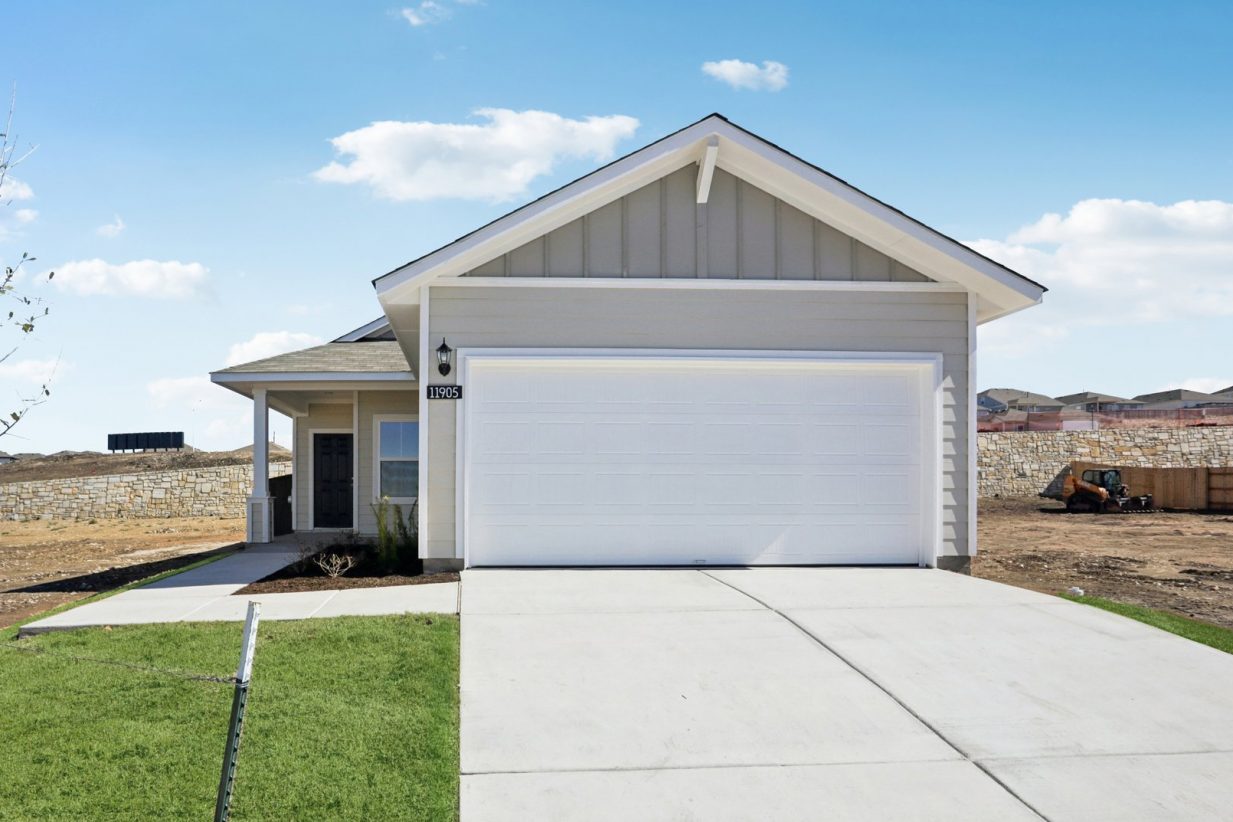 Image of a grey one story house with a white garage, a cement driveway, green grass and a blue sky in the background