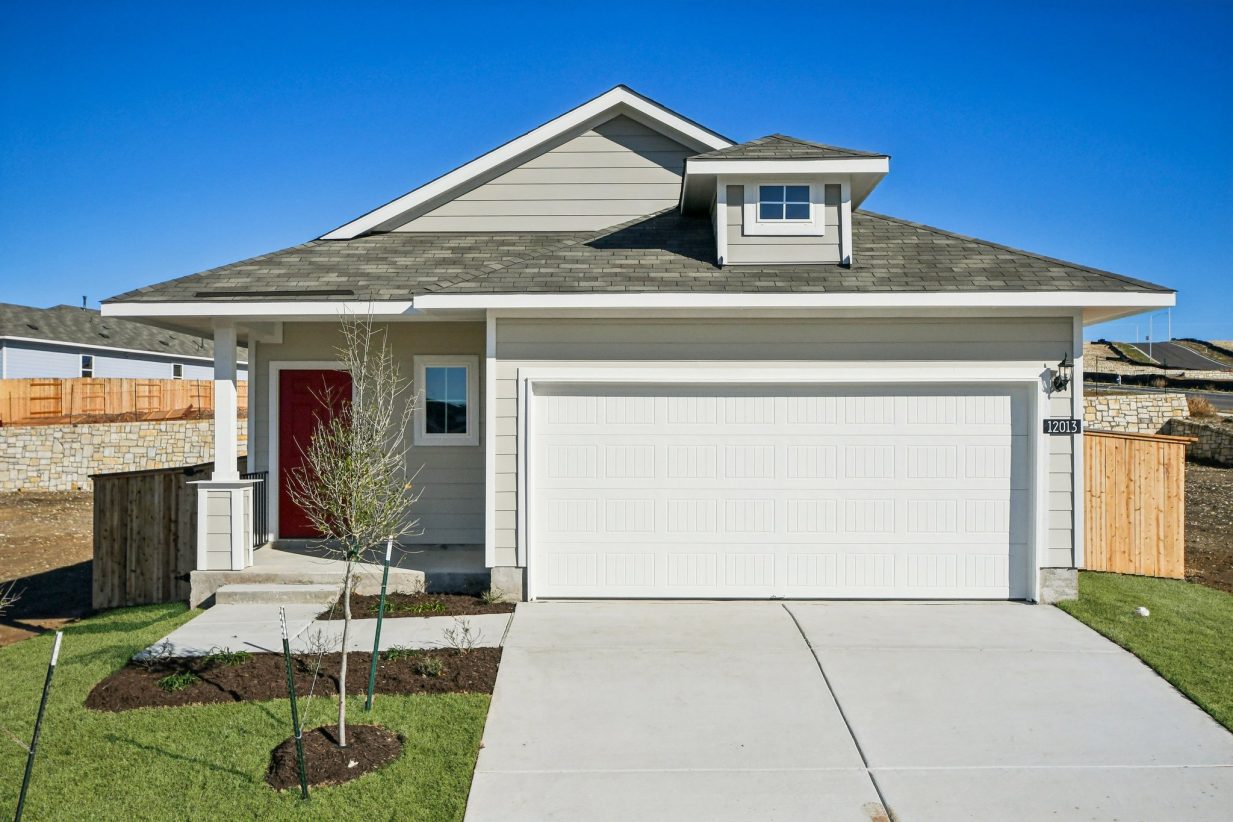 Image of a one story grey one story house with a red front door, white garage door, a driveway and a blue sky in the background