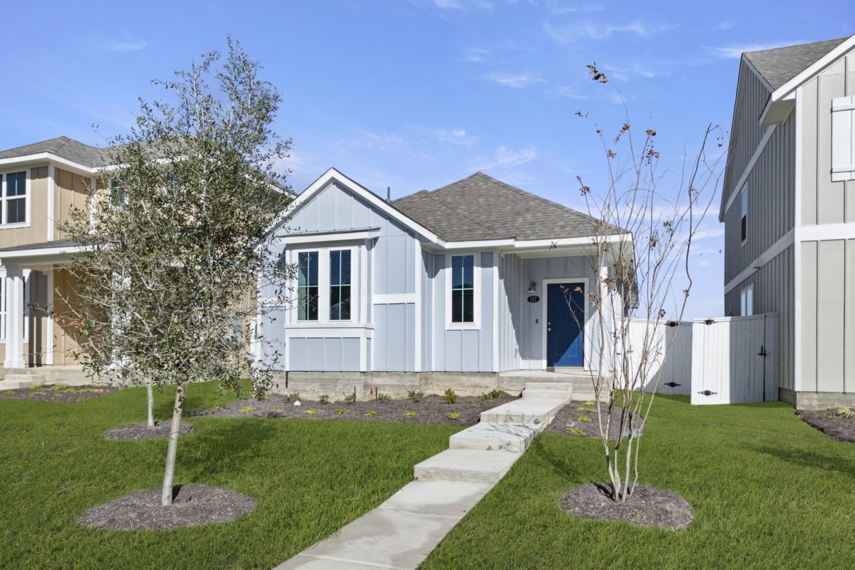 Image of the front exterior of a blue one story house with a royal blue door, a green grass front yard with a small tree and a blue sky in the background
