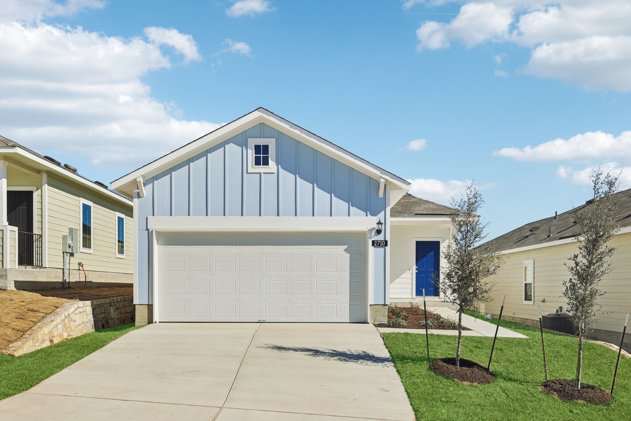Image of a blue one story home with a white garage door, royal blue front door, a cement driveway, a green grass front yard, and a blue sky in the background
