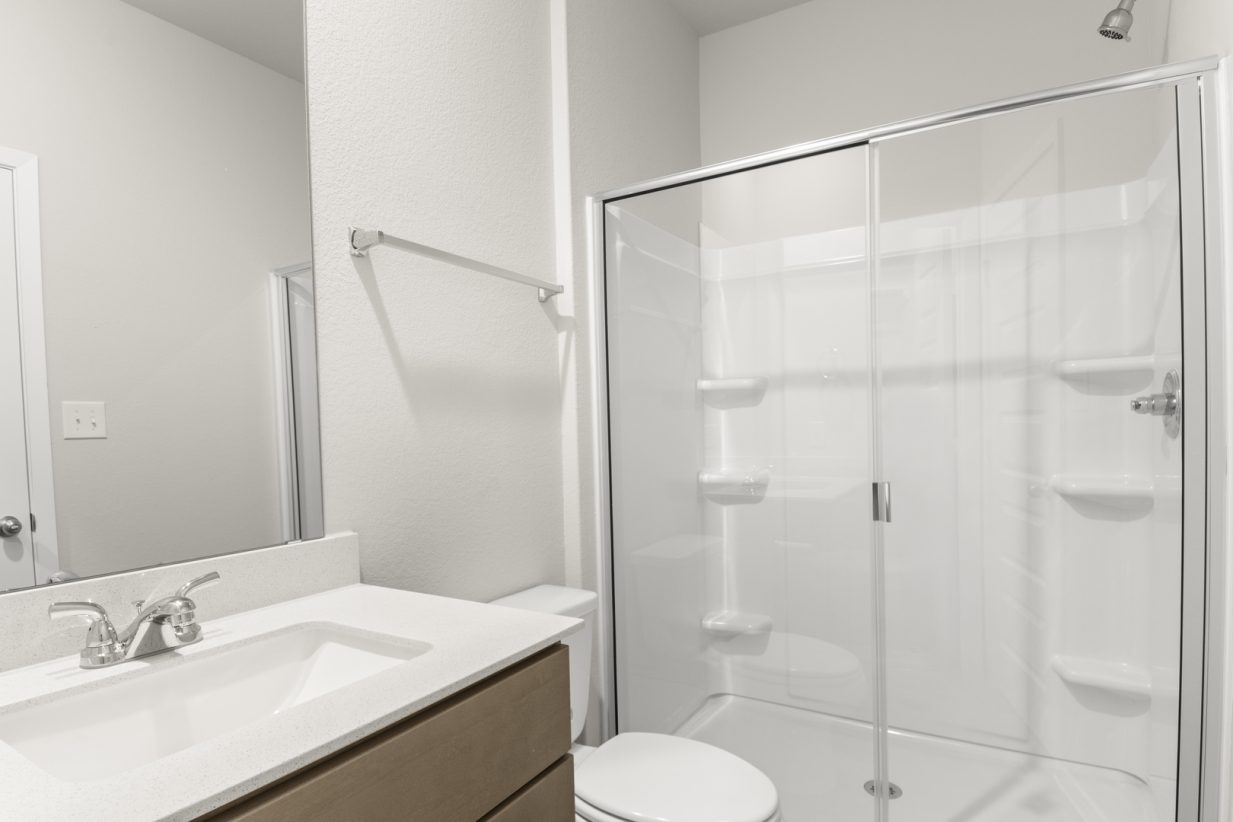 Image of a primary bathroom with a white vinyl white walk-in shower and a white vanity with brown cabinets