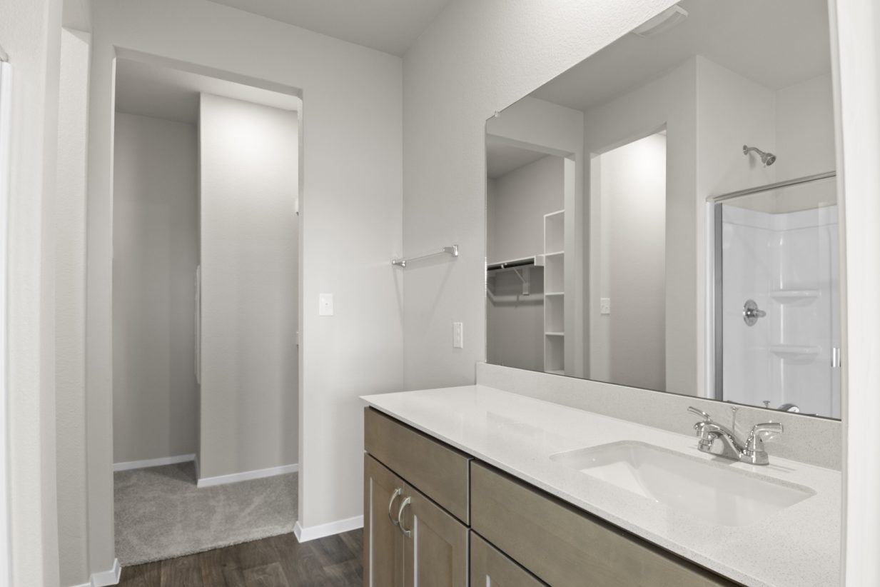 Image of a bathroom with a large mirror and white vanity with light brown cabinets with a walk-in shower reflected in the mirror. Open doorway to walk-in closet