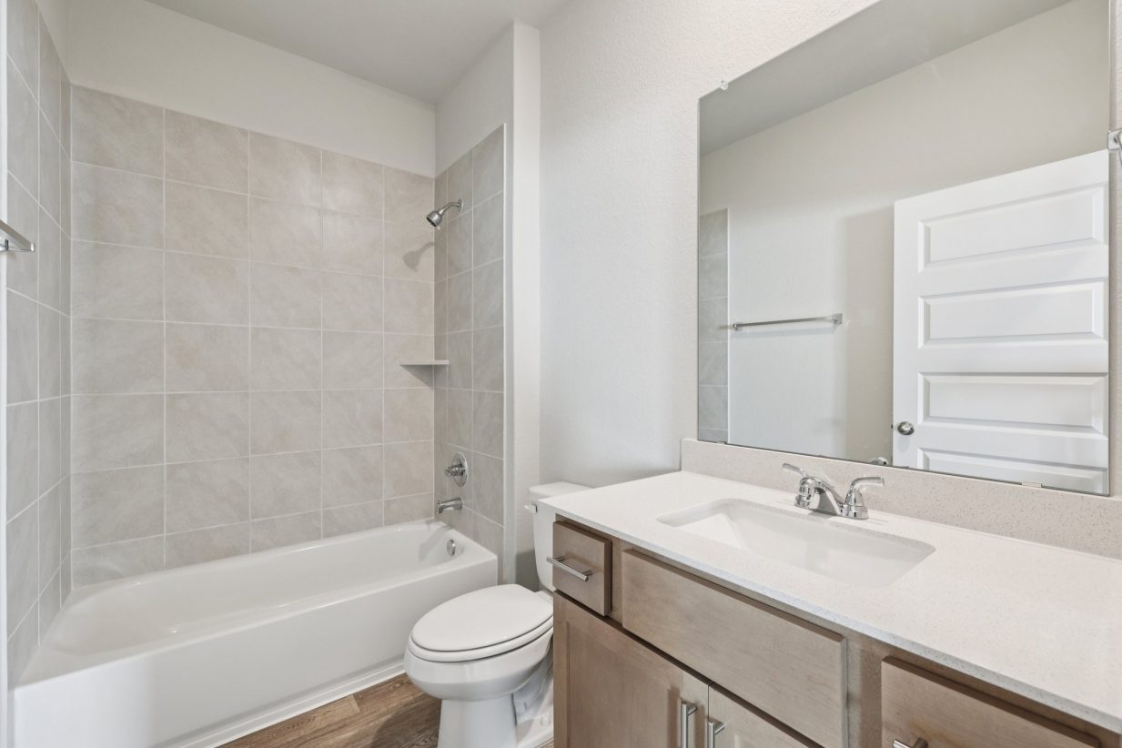 Image of a bathroom with a white dual vanity, brown cabinets, a large mirror, and a white tub with beige tiles