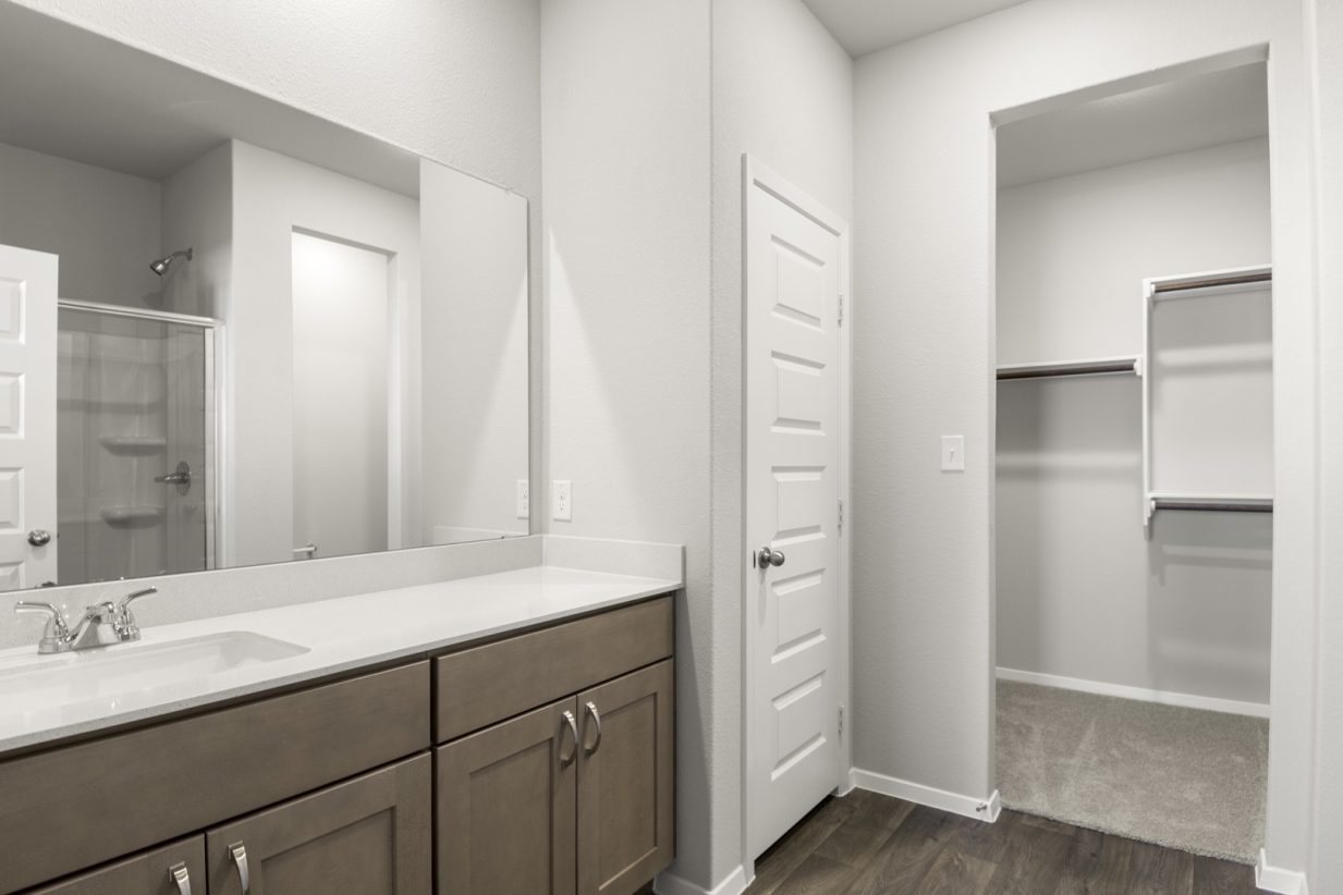 Image of a primary bathroom with a double white vanity, brown cabinets, a walk-in closet and a large mirror