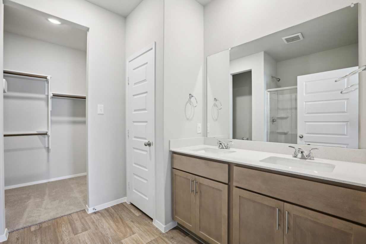 Image of a primary bathroom with light grey walls, brown cabinets, a white jack and jill vanity, a mirror and a walk-in closet