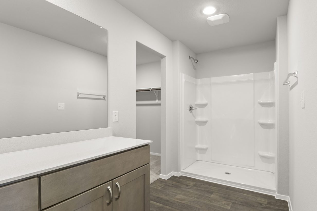 Image of a primary bathroom with brown cabinets, a white double vanity, a large mirror, a vinyl walk-in shower and a walk-in shower