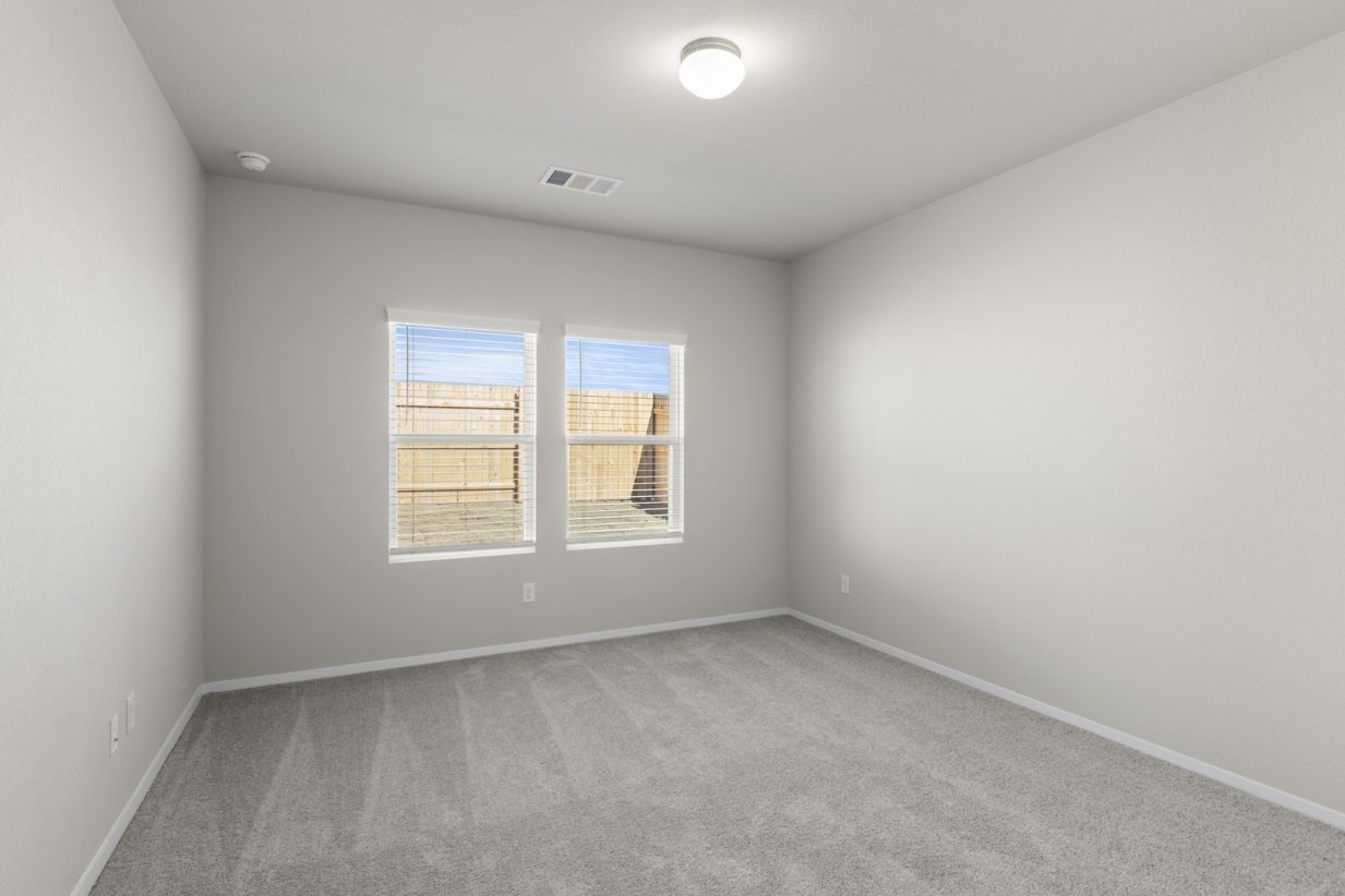 Image of a primary bedroom with beige walls, tan carpeting, two windows with blinds, and white trim