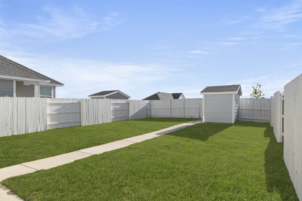Image of a backyard with a small grey detached shed, green grass, a white wooden fence and a cement pathway