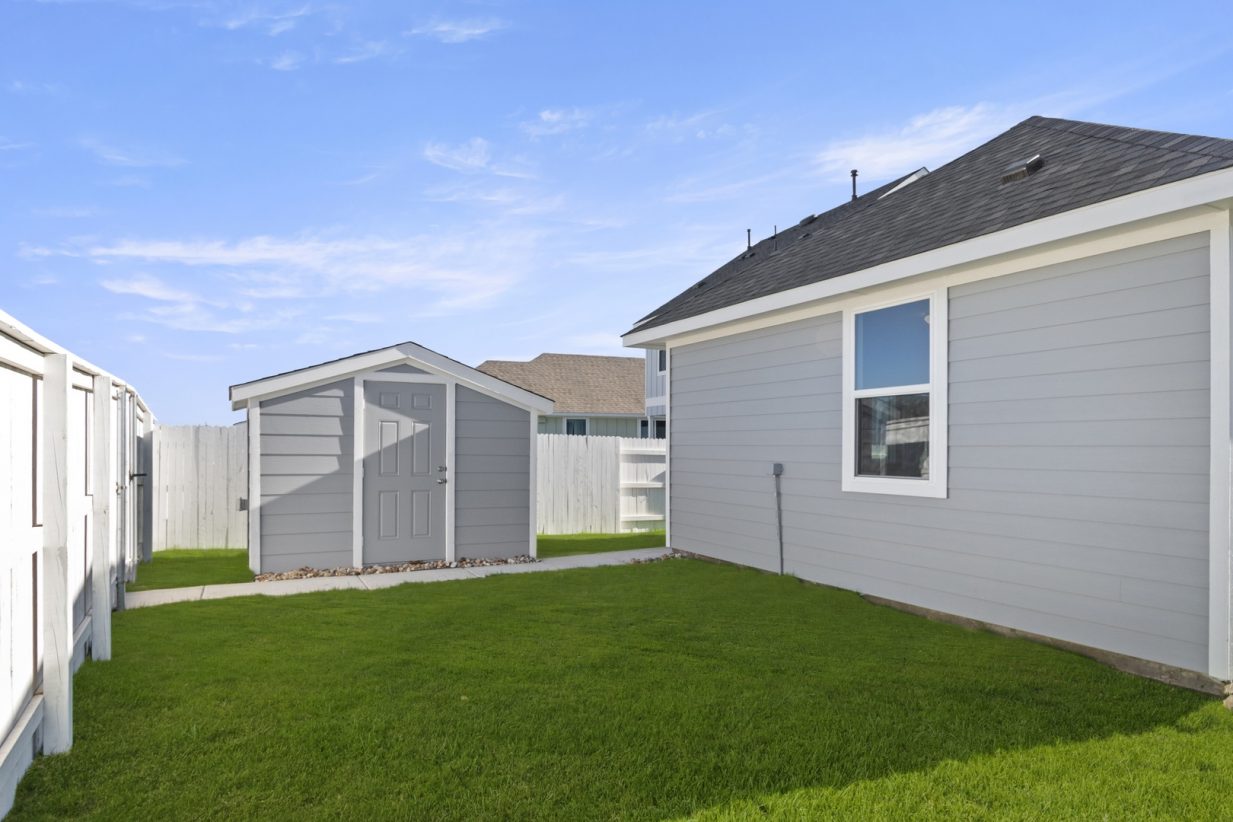 Image of a grey one story home backyard with a small grey shed, a white wooden fence, green grass and a blue sky