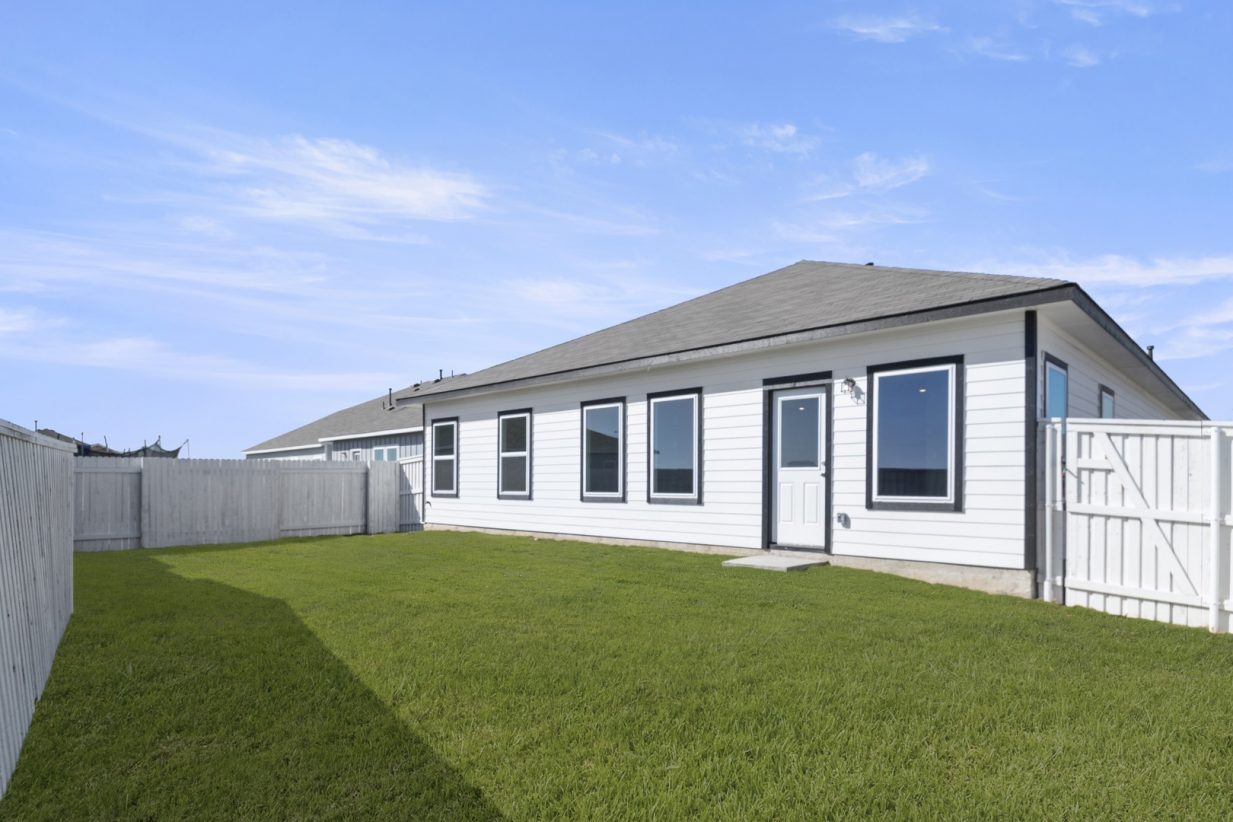 Image of the back exterior of a white one story house with black trim, windows, a white wooden fence, green grass and a blue sky in the background