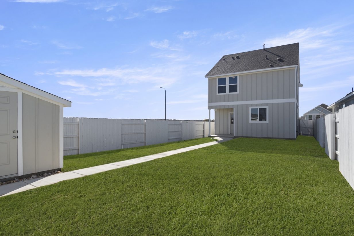 Image of a tan two story house back exterior with a green grass backyard, a small tan shed, a white fence and a blue sky