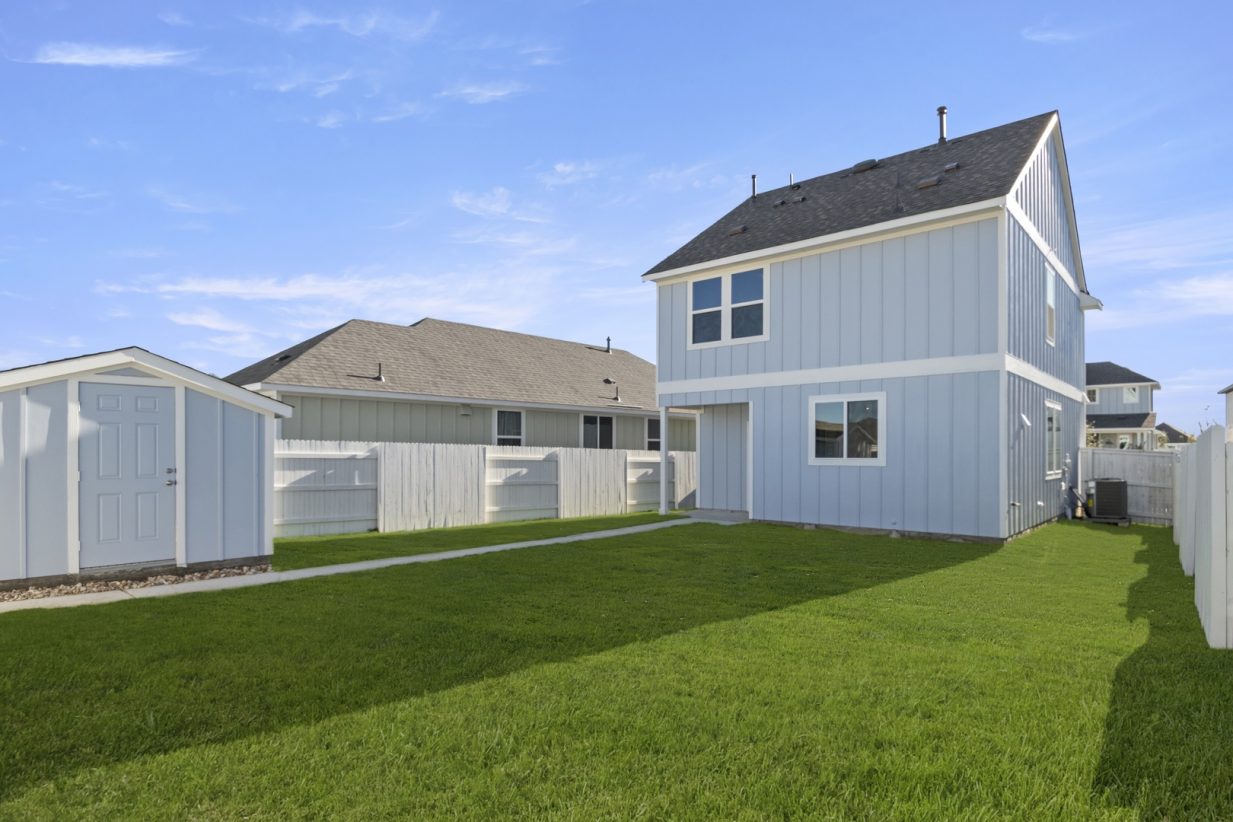 Image of a blue two story home back exterior with small blue detached shed, green grass, and a white wooden fence