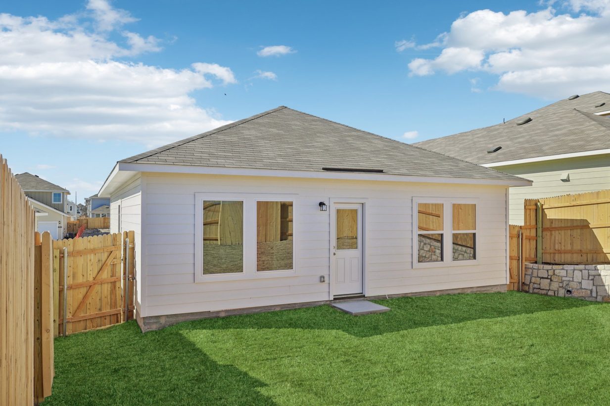 Image of a back exterior of a one story home with windows, a back door, a green grass back yard, a wooden fence and a blue sky in the background