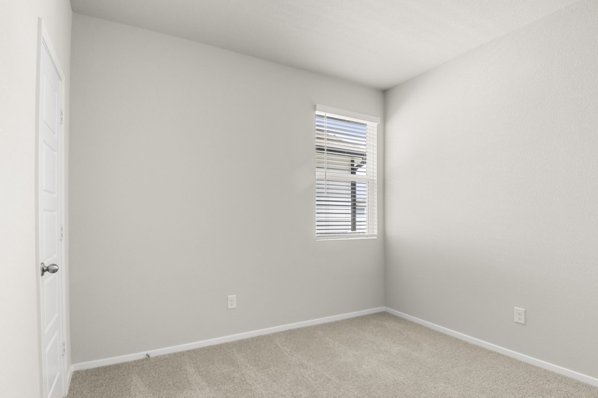 Image of a bedroom with tan carpeting and light grey walls with a window