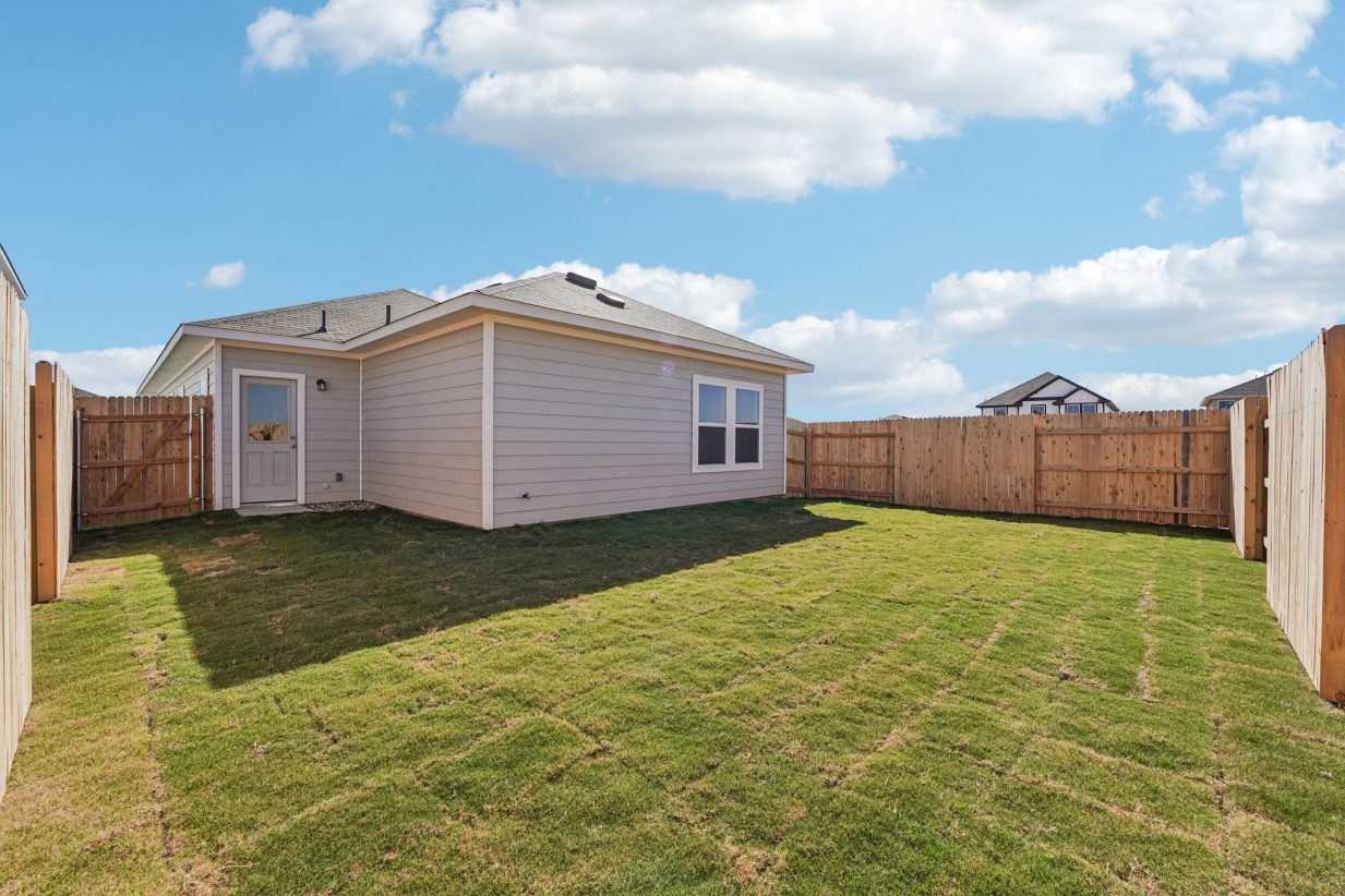 Image of a one story grey house with a green grass backyard, wooden fence and a blue sky in the background