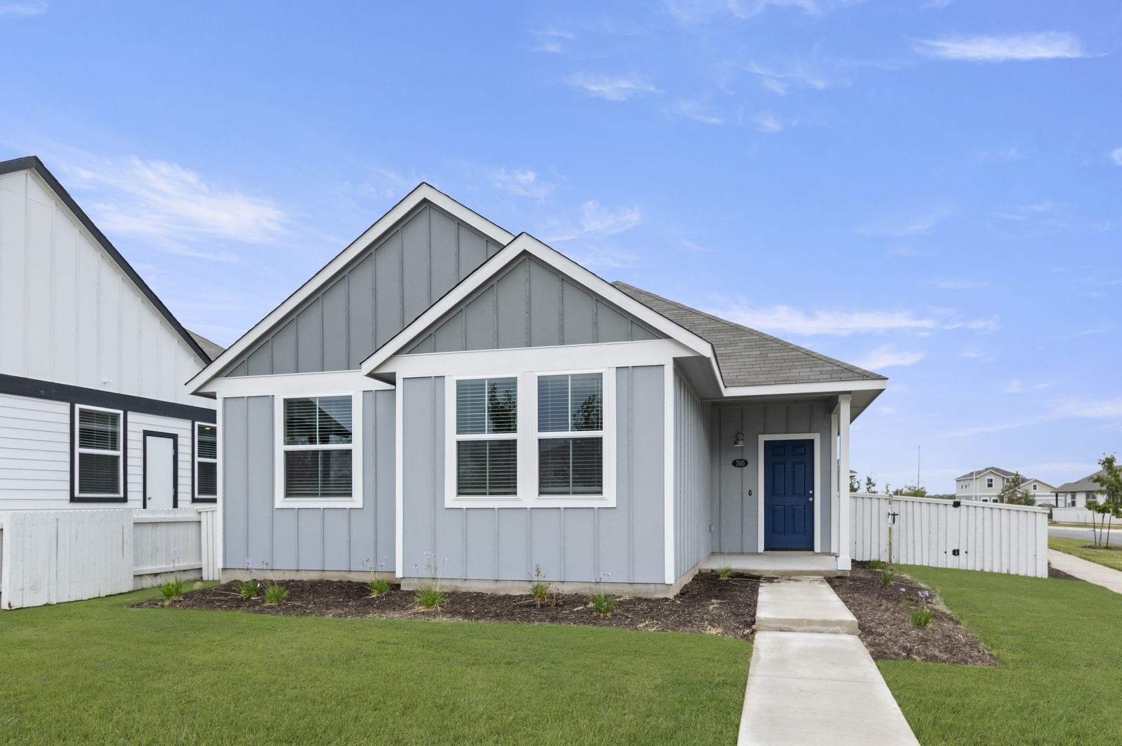Image of the front exterior of a grey one story home with white trim and a blue front door, a white fence, green grass, and a blue sky in the background