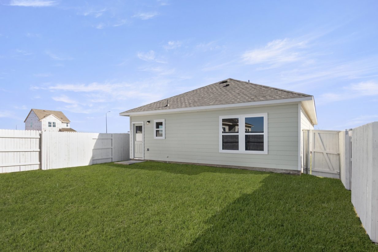 Image of a back exterior of a light green one-story house with white trim, a white fence and a green grass backyard