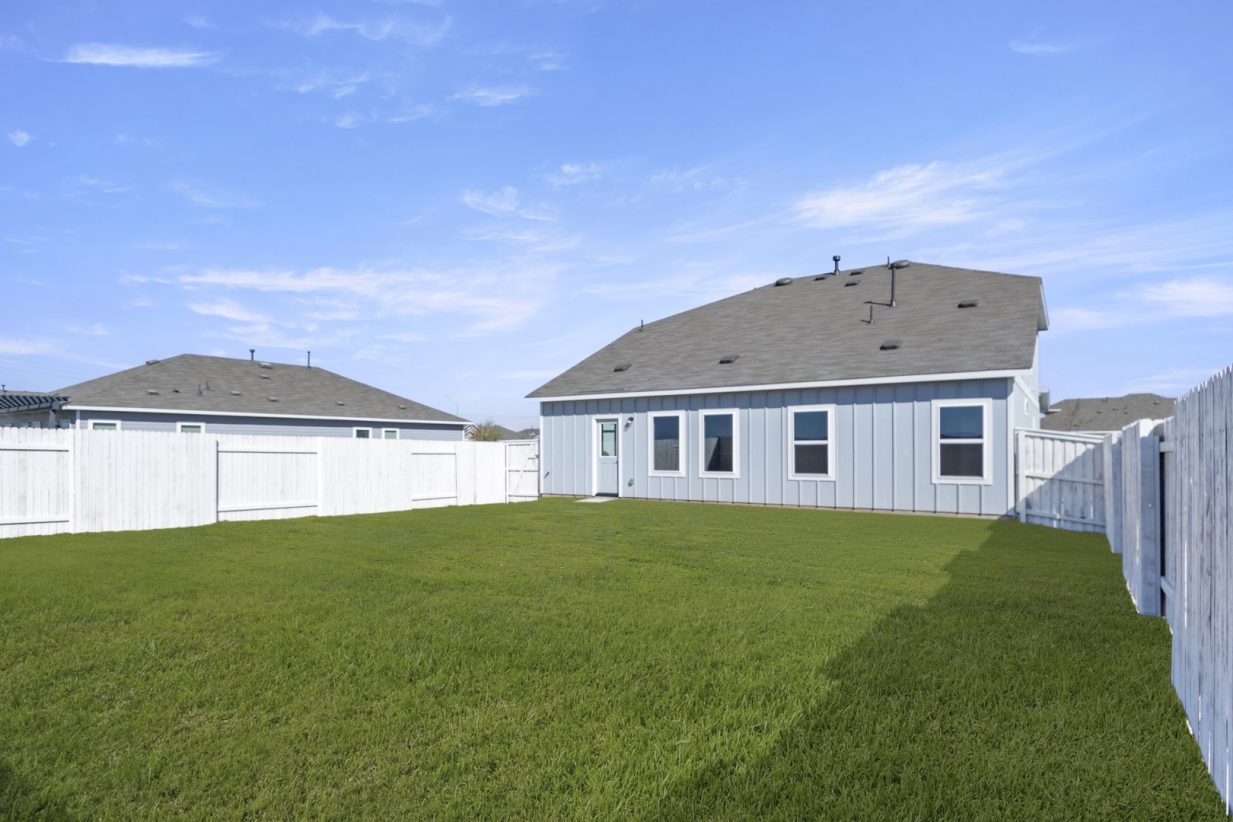 Image of a blue house back exterior with green grass backyard, a white wooden fence and a blue sky in the background