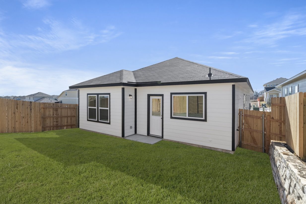 Image of a white one story house back exterior with black trim, a green grass backyard, a wooden fence and a blue sky in the background