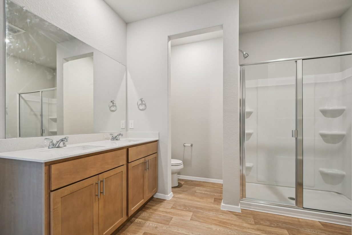 Image of a primary bathroom with light grey walls, brown cabinets, a large window and a walk-in shower
