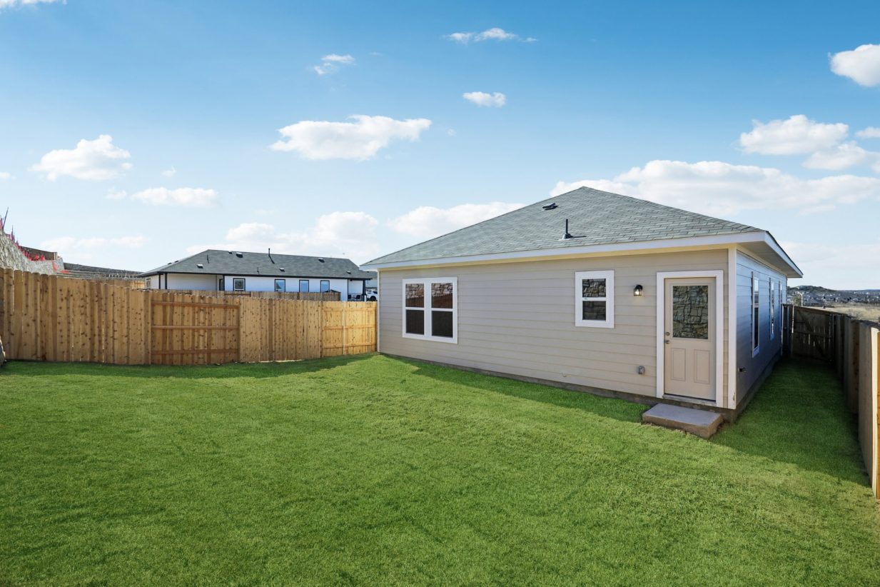 Image of a grey one story house with green grass, a wooden fence, and a blue sky in the background