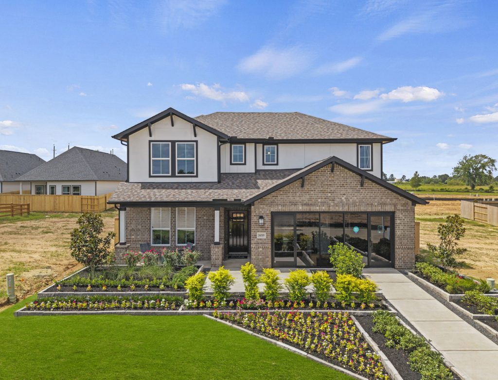Photo of two-story home with light paint, black accents, and dark brick on the first floor. Full wall windows in lieu of garage door. Walkway leading to the front door with green grass and landscaping around it
