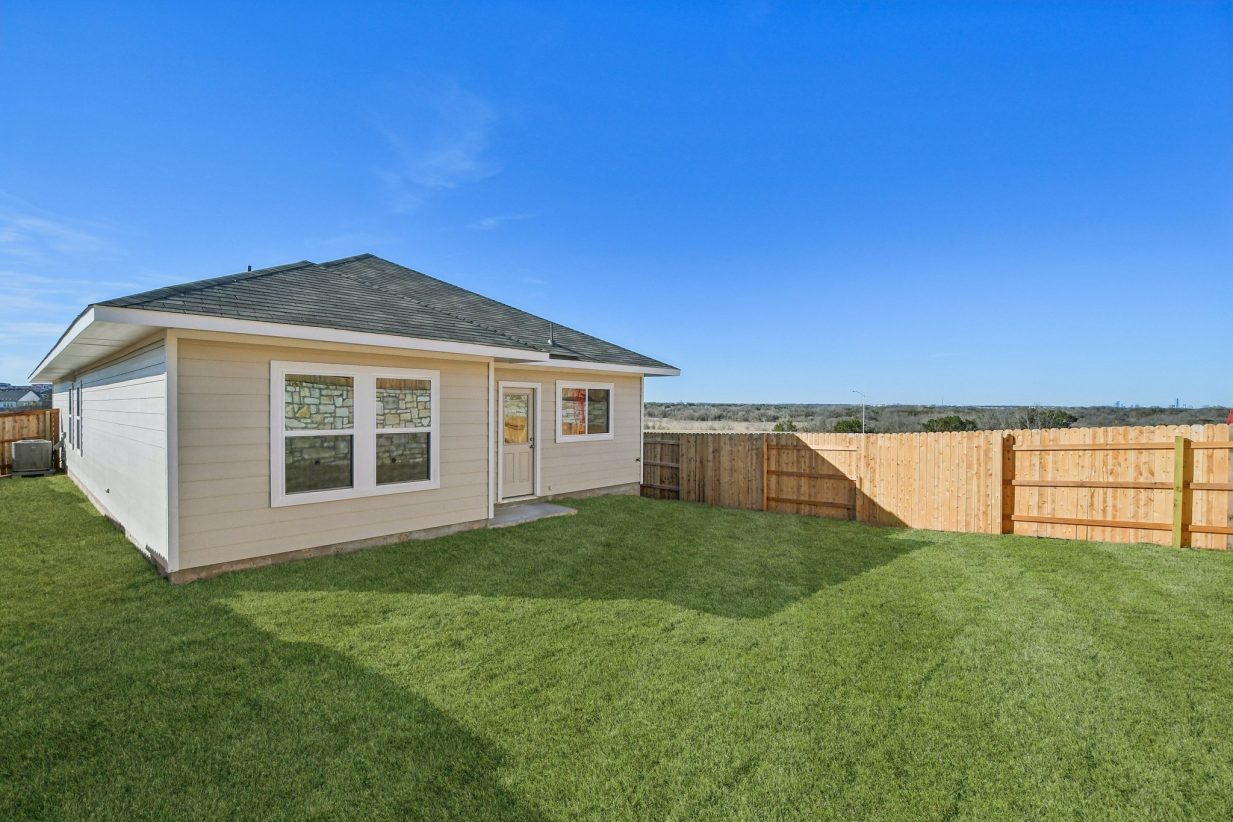 Image of a grey one story house back exterior with green grass, a wooden fence and a blue sky in the background