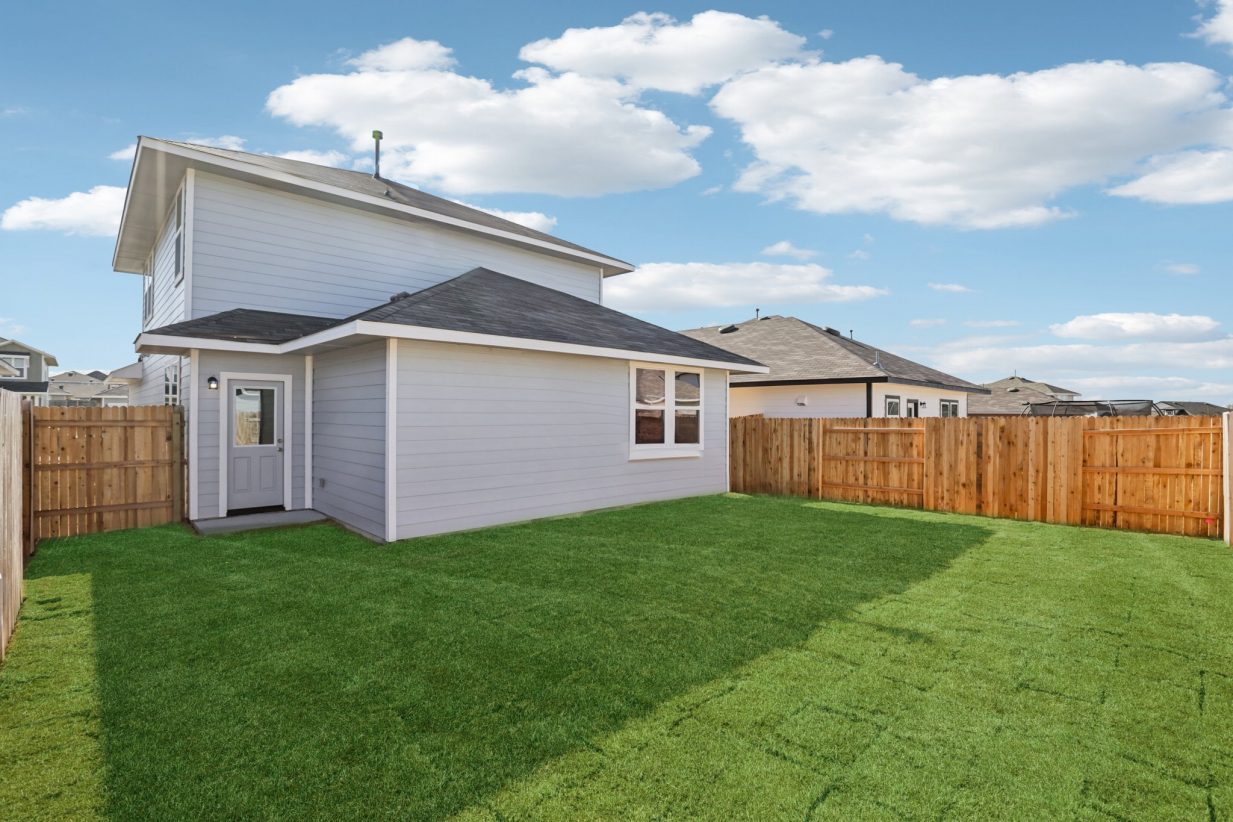 Image of the back exterior of a two story grey house with a green grass backyard, a wooden fence, and a blue sky in the background