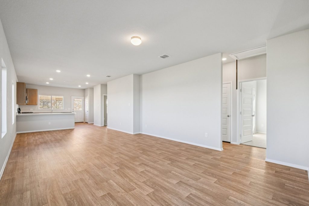 Image of a living room with light grey walls, vinyl flooring, and a kitchen in the distance